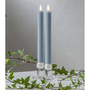 Kék LED vacsoragyertyák dekor pohárban, meleg fényű lánggal, természetes zöld díszítéssel és elegáns hangulatban elrendezve.