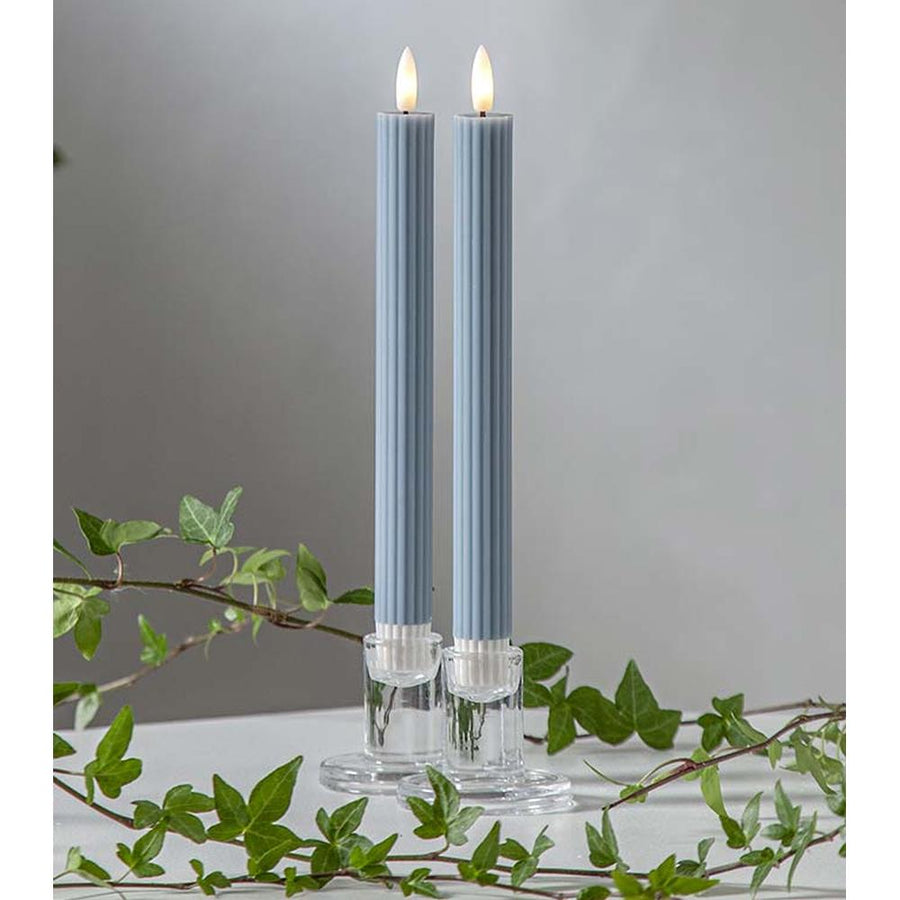 Kék LED vacsoragyertyák dekor pohárban, meleg fényű lánggal, természetes zöld díszítéssel és elegáns hangulatban elrendezve.