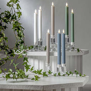 Pislákoló fényű LED vacsoragyertyák poharakban, hangulatos, természetes környezetben, zöld ágakkal körülvéve.