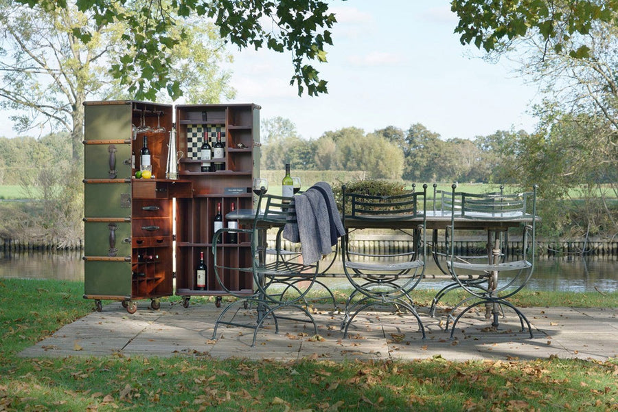 Kertben álló vintage stílusú zöld és mahagóni színű, görgős utazóláda-bárszekrény kertibútorokkal.