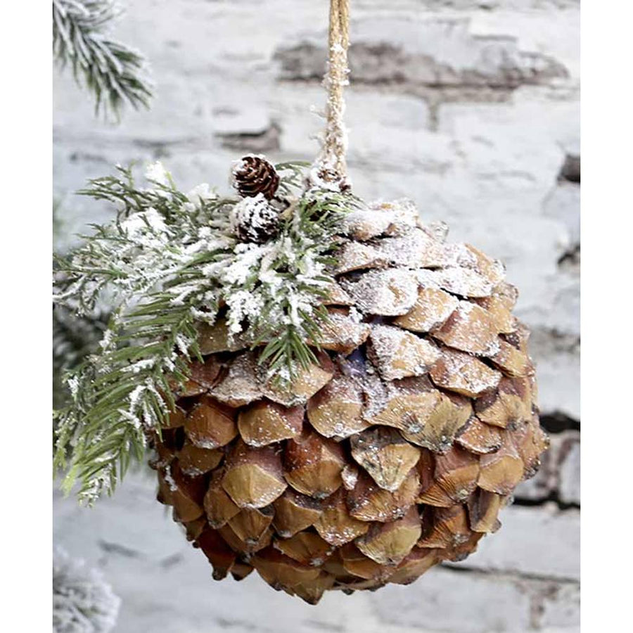 Vidéki stílusú, 15 cm átmérőjű, fenyőággal díszített, havas felületű, karácsonyi toboz gömb dísz, romos fal lelőtt.