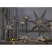 Különböző méretű, szürkeszínű karácsonyi papírcsillag lámpák, ajándékokkal és zöld fenyőágakkal..