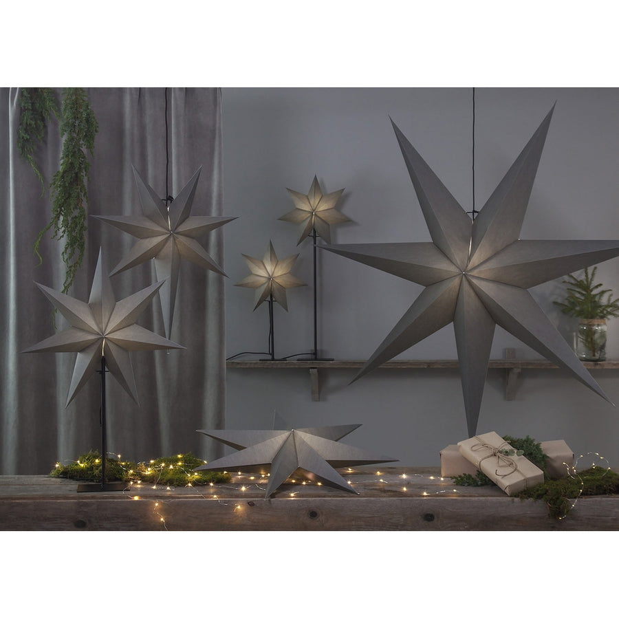 Különböző méretű, szürkeszínű karácsonyi papírcsillag lámpák, ajándékokkal és zöld fenyőágakkal..