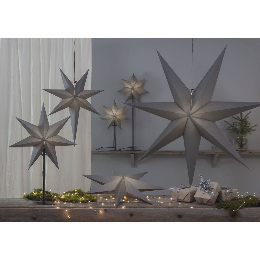 Különböző méretű, szürkeszínű karácsonyi papírcsillag lámpák, ajándékokkal és zöld fenyőágakkal.