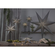 Különböző méretű, szürkeszínű karácsonyi papírcsillag lámpák, ajándékokkal és zöld fenyőágakkal..