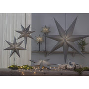 Különböző méretű, szürkeszínű karácsonyi papírcsillag lámpák, ajándékokkal és zöld fenyőágakkal..