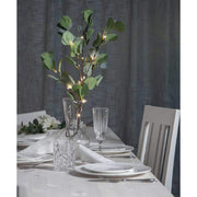 Élethű, 90 cm magas, mesterséges eukaliptusz ág műnövény, beépített, időzíthető meleg fehér fényű, harmatcsepp formájú LED világítással, ünnepi fehér asztalon, szürke háttér előtt , üveg borospoharakkal és  fehér színű terítékkel 