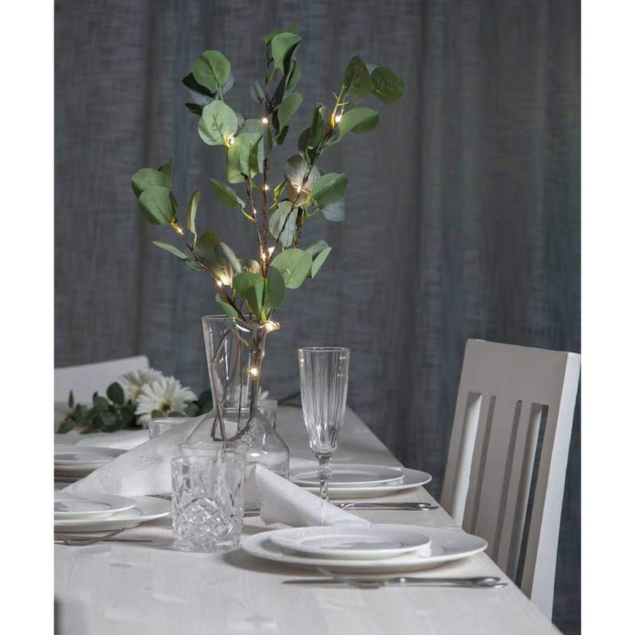 Élethű, 90 cm magas, mesterséges eukaliptusz ág műnövény, beépített, időzíthető meleg fehér fényű, harmatcsepp formájú LED világítással, ünnepi fehér asztalon, szürke háttér előtt , üveg borospoharakkal és  fehér színű terítékkel 