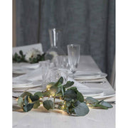 Élethű, 90 cm magas, mesterséges eukaliptusz ág műnövény, beépített, időzíthető meleg fehér fényű, harmatcsepp formájú LED világítással, ünnepi fehér asztalon, szürke háttér előtt , üveg borospoharakkal és fehér színű terítékkel