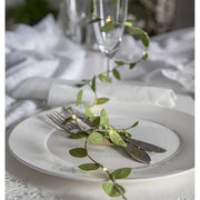 Mesterséges, 95 cm hosszú apró, zöld levelű girland műnövény, meleg fehér fényű, harmatcsepp formájú LED világítással ünnepi fehér terített asztalon