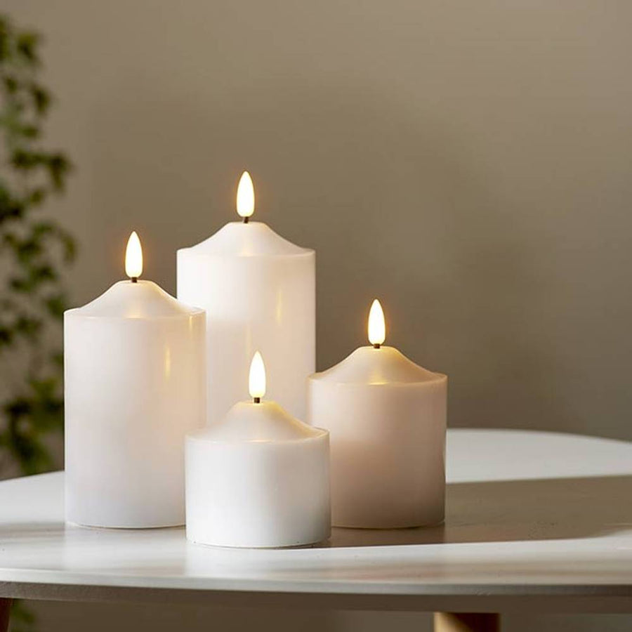 4 db különböző méretű, fehér színű viaszból készült led gyertya, meleg fehér fényű led lángokkal, fehér modern asztalon, szürke fal előtt