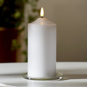 Valódi viaszból készült, fehér színű led gyertya meleg fehér fenyő led lánggal üveg tányéron, fehér színű asztalon, szürke fal előtt 