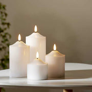4 db különböző méretű, fehér színű viaszból készült led gyertya, meleg fehér fényű led lángokkal,  fehér modern asztalon, szürke fal előtt