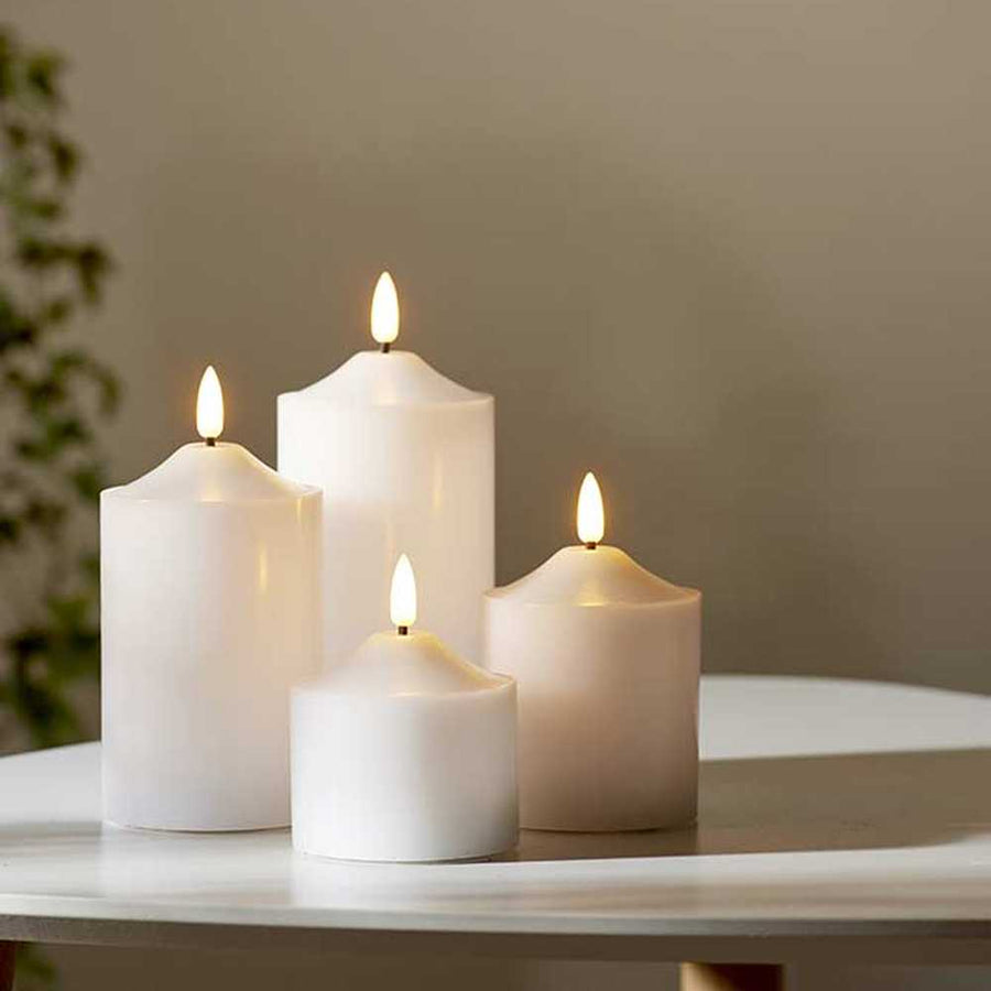 4 db különböző méretű, fehér színű viaszból készült led gyertya, meleg fehér fényű led lángokkal,  fehér modern asztalon, szürke fal előtt