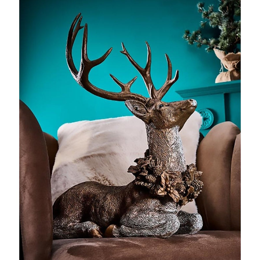 Erdei fenyőtoboz koszorúval díszített, nagyméretű karácsonyi szarvas figura , barna színű fotelbe, párnával.