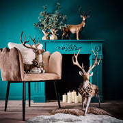 Erdei fenyőtoboz koszorúval díszített, nagyméretű karácsonyi szarvas figura, kék színű szobában, kék színű kandalló előtt.