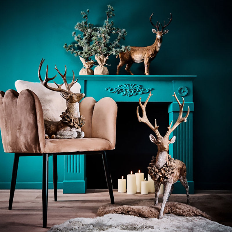 Erdei fenyőtoboz koszorúval díszített, nagyméretű karácsonyi szarvas figura, kék színű szobában, kék színű kandalló előtt.