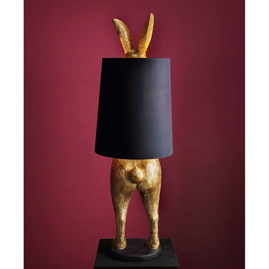 Aranyszínű, rejtőzködő nyuszi figurás, díjnyertes dizájn állólámpa, feketeszínű lámpaernyővel, bordó fal előtt.