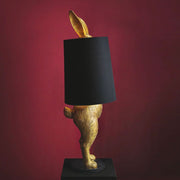 Aranyszínű, rejtőzködő nyuszi figurás, díjnyertes dizájn állólámpa, feketeszínű lámpaernyővel, bordó fal előtt.