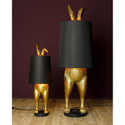 Aranyszínű, rejtőzködő nyuszi figurás, díjnyertes dizájn állólámpa, feketeszínű lámpaernyővel, bordó fal előtt.