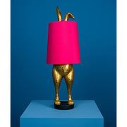 Aranyszínű, rejtőzködő nyuszi figurás, 74 cm magas, kortárs, glamour stílusú, díjnyertes dizájn asztali lámpa, pink színű lámpaernyővel, kékszínű háttér előtt.