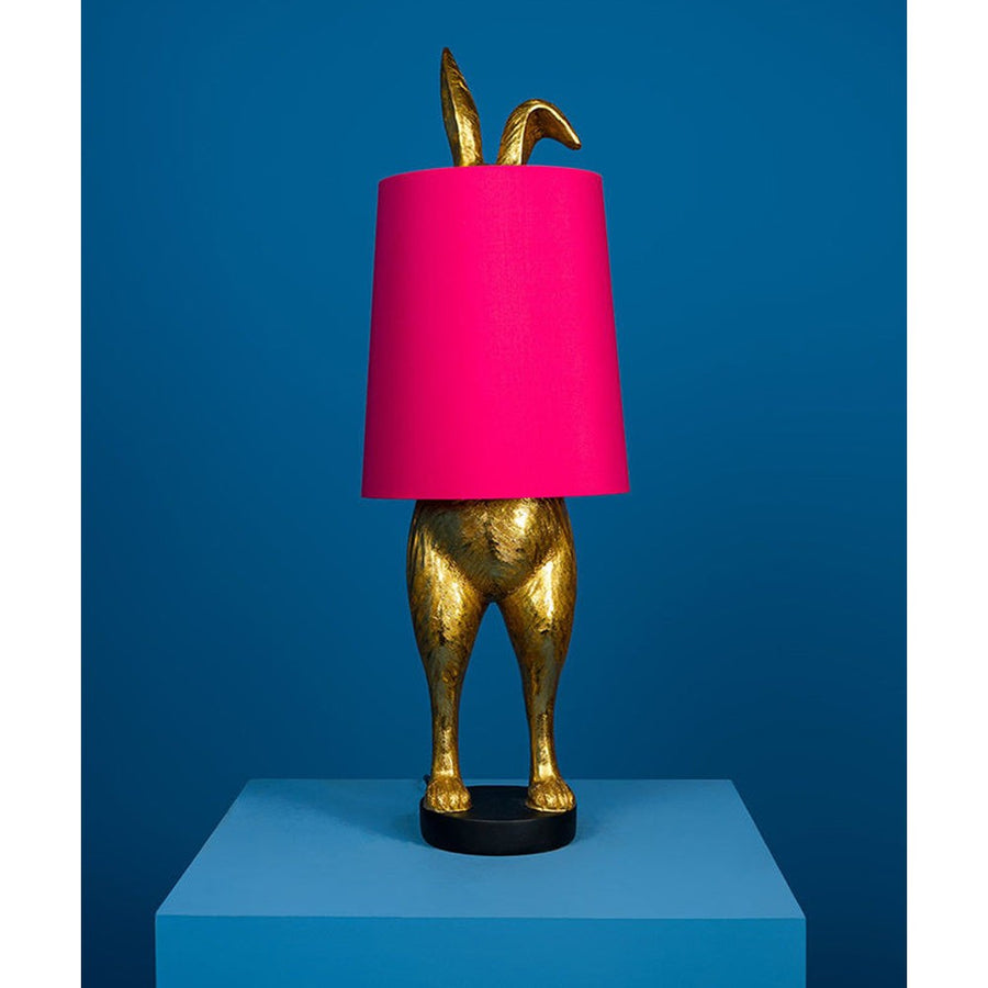 Aranyszínű, rejtőzködő nyuszi figurás, 74 cm magas, kortárs, glamour stílusú, díjnyertes dizájn asztali lámpa, pink színű lámpaernyővel, kékszínű háttér előtt.