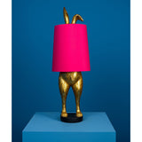 Aranyszínű, rejtőzködő nyuszi figurás, 74 cm magas, kortárs, glamour stílusú, díjnyertes dizájn asztali lámpa, pink színű lámpaernyővel, kékszínű háttér előtt.