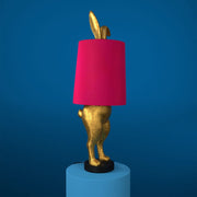 Aranyszínű, rejtőzködő nyuszi figurás, 74 cm magas, kortárs, glamour stílusú, díjnyertes dizájn asztali lámpa, pink színű lámpaernyővel, kékszínű háttér előtt.