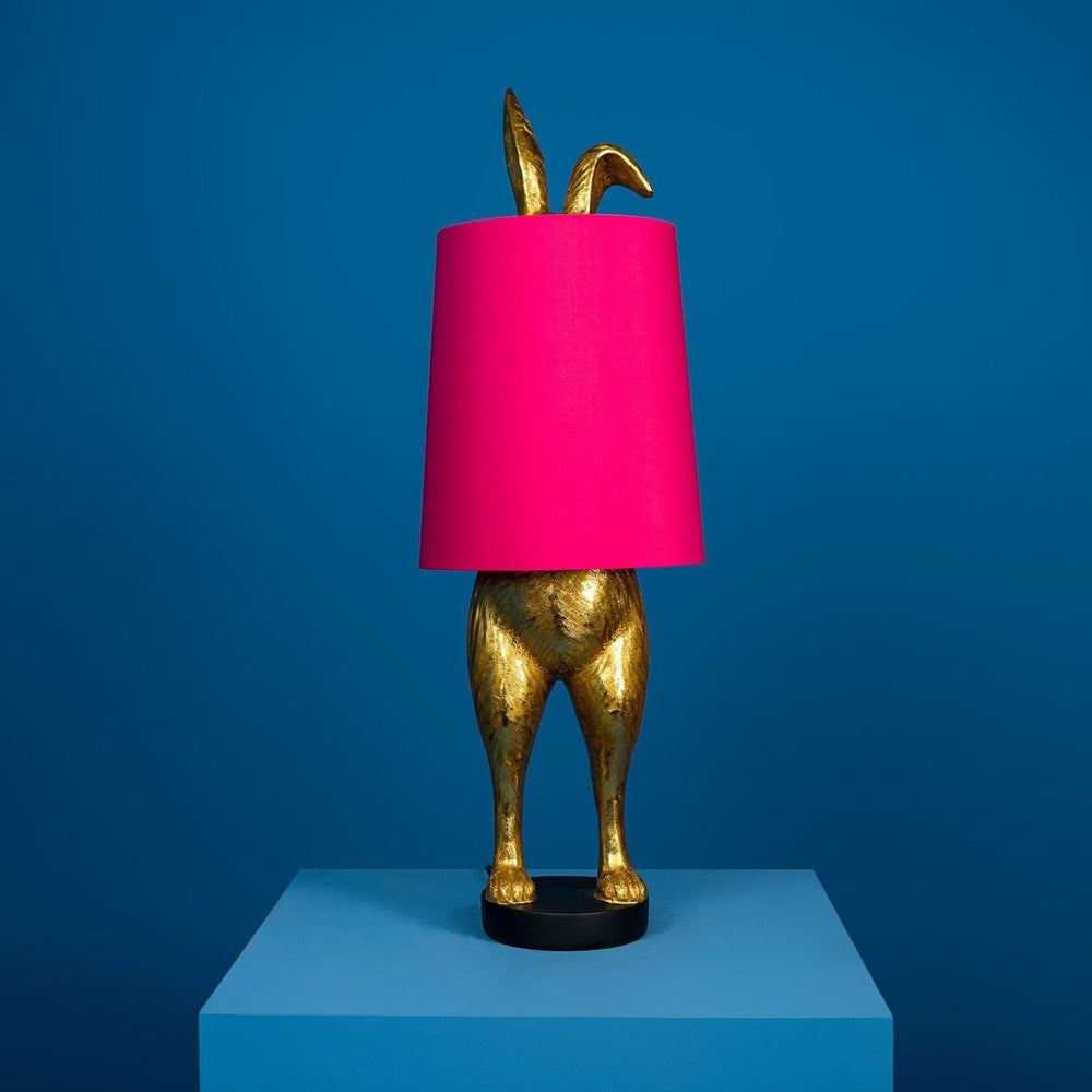 Aranyszínű, rejtőzködő nyuszi figurás, 74 cm magas, kortárs, glamour stílusú, díjnyertes dizájn asztali lámpa, pink színű lámpaernyővel, kékszínű háttér előtt.