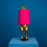 Aranyszínű, rejtőzködő nyuszi figurás, 74 cm magas, kortárs, glamour stílusú, díjnyertes dizájn asztali lámpa, pink színű lámpaernyővel, kékszínű háttér előtt.