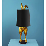 Aranyszínű, rejtőzködő nyuszi figurás, 74 cm magas, kortárs, glamour stílusú, díjnyertes dizájn asztali lámpa, feketeszínű lámpaernyővel, kék háttér előtt.