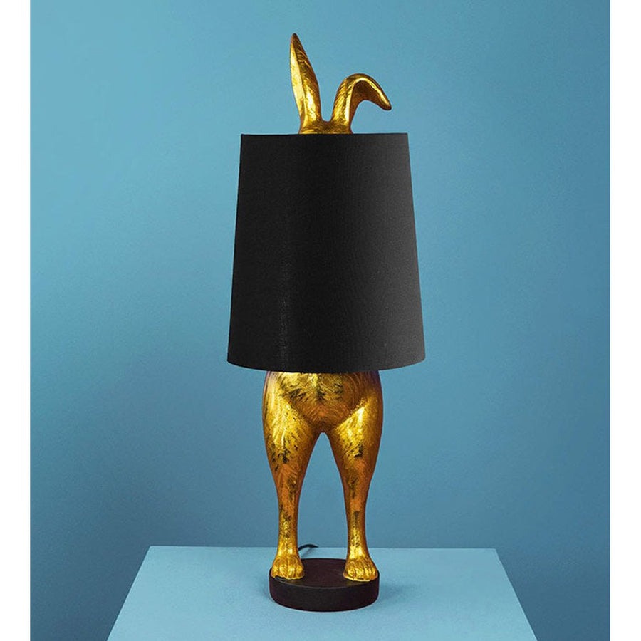 Aranyszínű, rejtőzködő nyuszi figurás, 74 cm magas, kortárs, glamour stílusú, díjnyertes dizájn asztali lámpa, feketeszínű lámpaernyővel, kék háttér előtt.