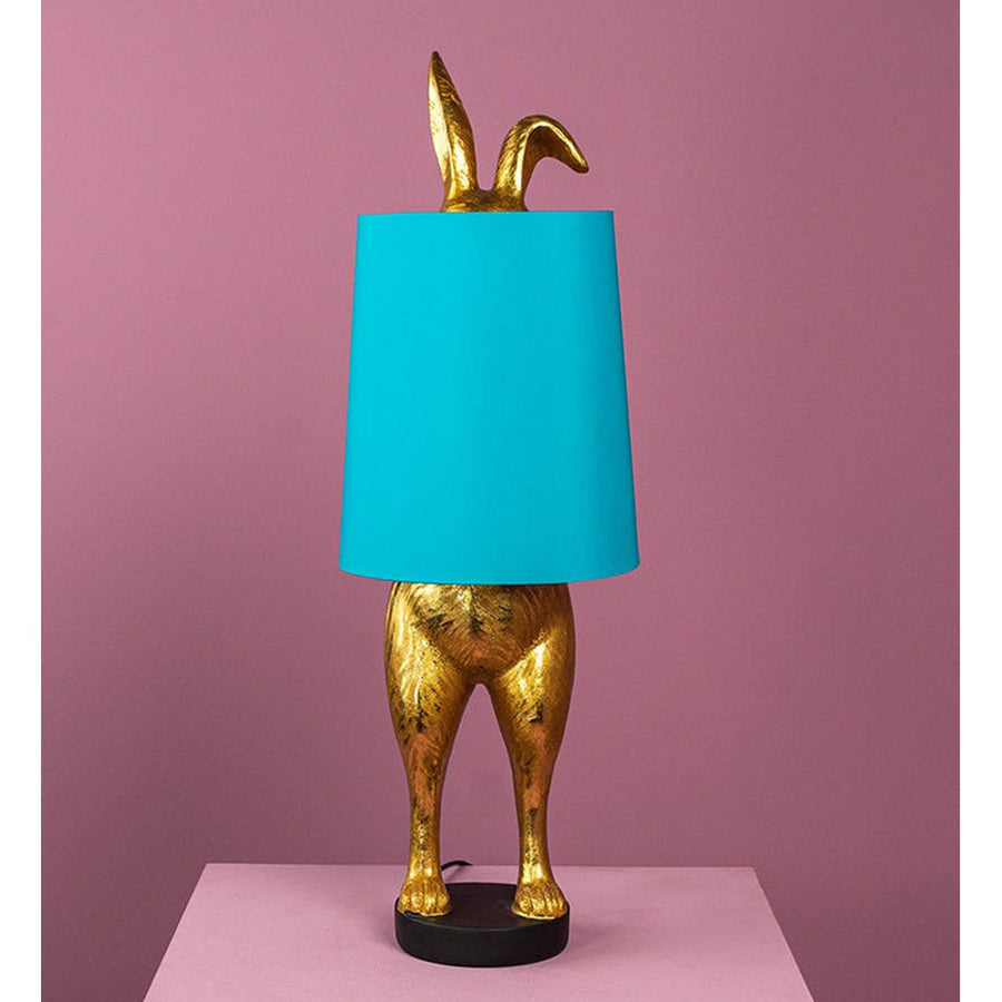 Aranyszínű, rejtőzködő nyuszi figurás, 74 cm magas, kortárs, glamour stílusú, díjnyertes dizájn asztali lámpa, türkizszínű lámpaernyővel, mályva színű fal előtt.
