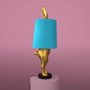 Aranyszínű, rejtőzködő nyuszi figurás, 74 cm magas, kortárs, glamour stílusú, díjnyertes dizájn asztali lámpa, türkizszínű lámpaernyővel, mályva színű fal előtt.