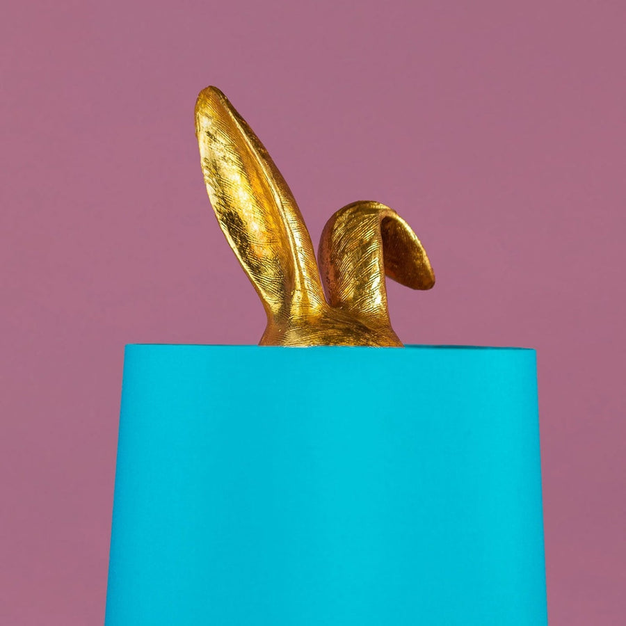 Aranyszínű, rejtőzködő nyuszi figurás, 74 cm magas, kortárs, glamour stílusú, díjnyertes dizájn asztali lámpa, türkizszínű lámpaernyővel, mályva színű fal előtt.