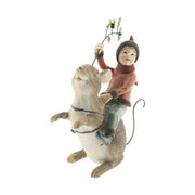 Barna színű erdei egér hátán utazó karácsonyi manó bordó ruhában és sállal, kezében magyallal szemből fotózott képe