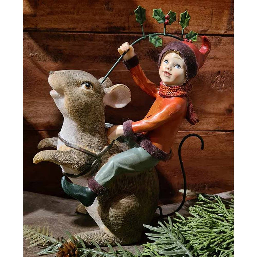 Barna színű erdei egér hátán utazó karácsonyi manó bordó ruhában és sállal, kezében magyallal barna színű deszkafal előtt 