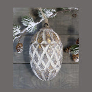 Vintage stílusú, mangófából faragott, 15 cm magas, antikolt felületű óarany színű, fenyőtoboz formájú, kézműves karácsonyfadísz szürke deszka asztalon havas fenyővel 