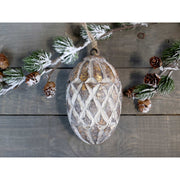 Vintage stílusú, mangófából faragott, 15 cm magas, antikolt felületű óarany színű, fenyőtoboz formájú, kézműves karácsonyfadísz szürke deszka asztalon havas fenyővel.