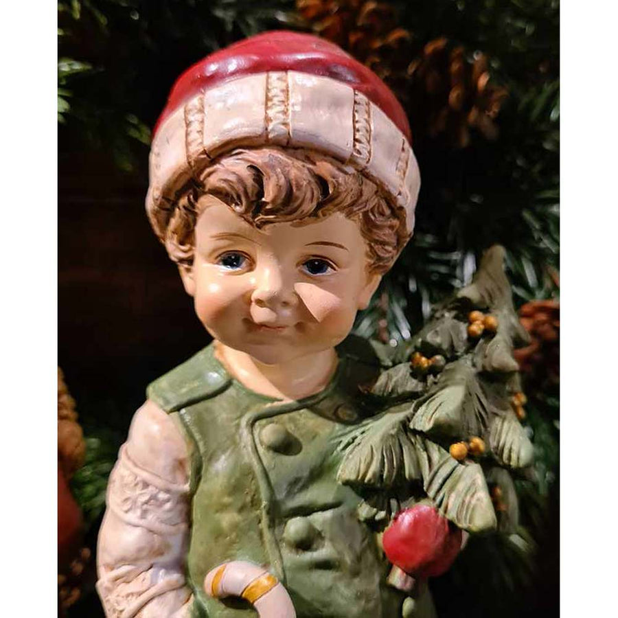 Vintage stílusú, 24,2 cm magas, tradicionális megjelenésű, karácsonyi téli sapkás kisfiú karácsonyfával a kezében zöld fenyőfa előtt