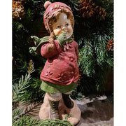 Vintage stílusú, 24,7 cm magas, tradicionális megjelenésű, karácsonyi téli sapkás kislány sállal a nyakában zöld fenyő előtt 