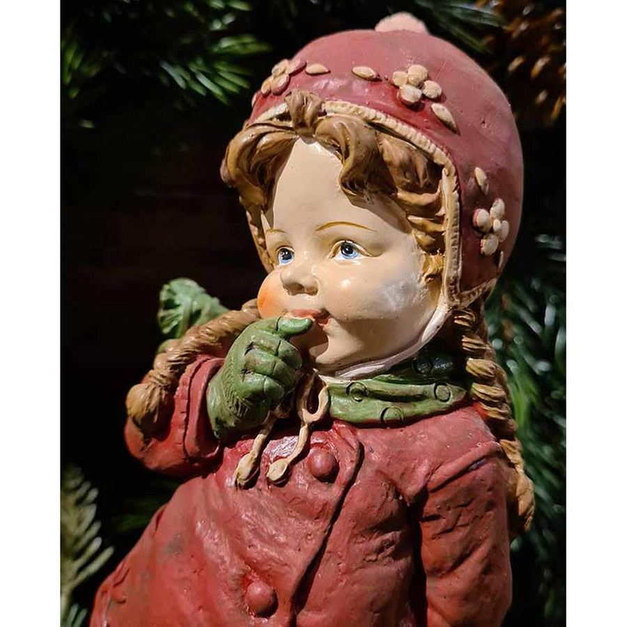 Vintage stílusú, 24,7 cm magas, tradicionális megjelenésű, karácsonyi téli sapkás kislány sállal a nyakában zöld fenyő előtt