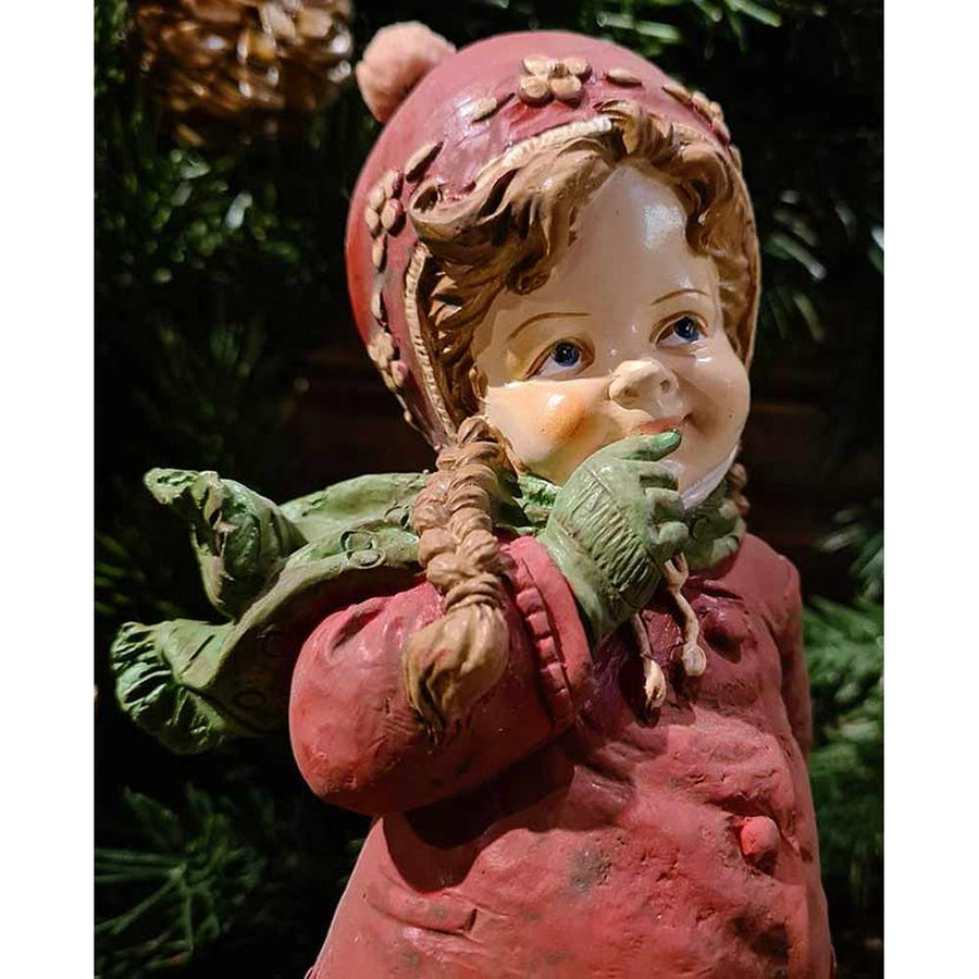 Vintage stílusú, 24,7 cm magas, tradicionális megjelenésű, karácsonyi téli sapkás kislány sállal a nyakában zöld fenyő előtt